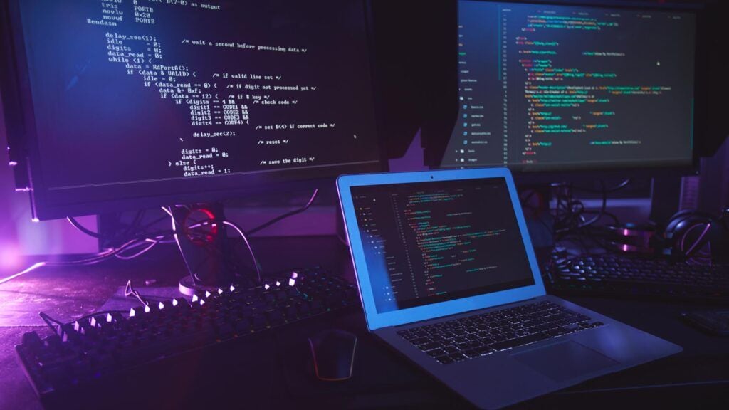 Hintergrundbild von verschiedenen Computergeräten mit Programmiercode auf Bildschirmen auf einem Tisch in einem dunklen Raum, Cybersicherheitskonzept, Kopierraum.