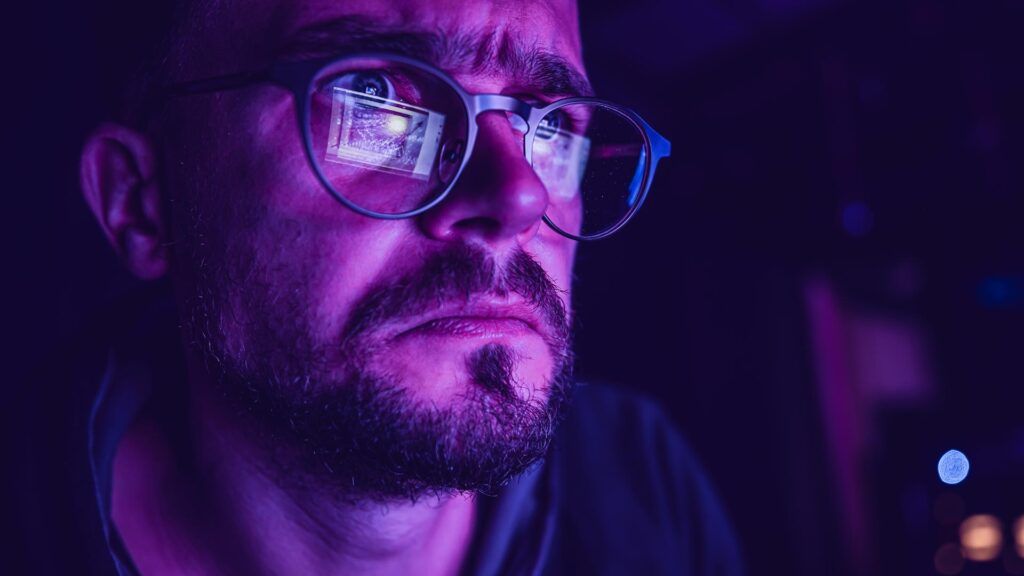 A man wearing glasses stares intently at a computer screen late in the evening.