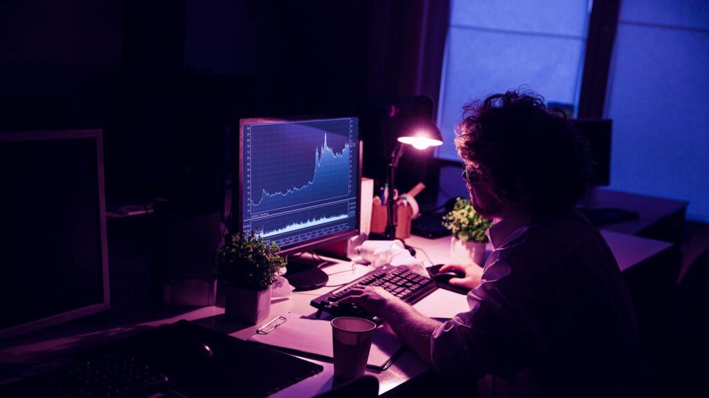 Work with diagrams. Man works in office alone during COVID-19 quarantine, staying late at night. Young businessman, manager performs tasks with smartphone, laptop, tablet in empty workspace.